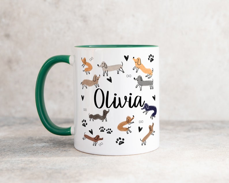 Pode incluir: Uma caneca de cer&acirc;mica branca com uma asa e rebordo interior verde-escuro. O exterior apresenta um padr&atilde;o repetitivo de c&atilde;es dachshund de v&aacute;rias cores, pegadas pretas, pequenos cora&ccedil;&otilde;es e ossos, com o nome Olivia em letra preta.