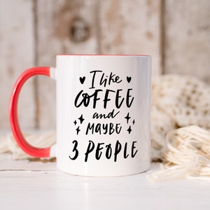 May include: White ceramic coffee mug with a red rim and handle. The mug has black text that reads "I like coffee and maybe 3 people".