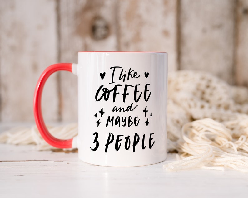 May include: White ceramic coffee mug with a red rim and handle. The mug has black text that reads "I like coffee and maybe 3 people".