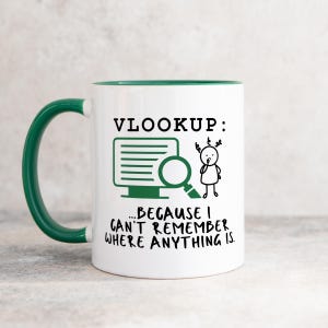 May include: White coffee mug with a green rim and handle. The mug has a green cartoon illustration of a person looking at a computer screen with a magnifying glass. The text on the mug reads "VLOOKUP: ... BECAUSE I CAN'T REMEMBER WHERE ANYTHING IS."