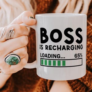 Può includere: Una tazza in ceramica bianca con testo nero che recita BOSS IS RECHARGING e LOADING... 65% insieme a una barra di caricamento verde. La tazza presenta un manico standard e un corpo cilindrico liscio, mostrando il design stampato sul davanti.