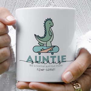 May include: White ceramic mug featuring a green dinosaur riding a skateboard. The text reads "Auntie-saurus like a normal aunt but more rawr-some!"