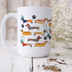 May include: White ceramic coffee mug with a colorful illustration of dachshunds wearing different costumes and accessories, including astronaut helmets, hot dog costumes, and roller skates.