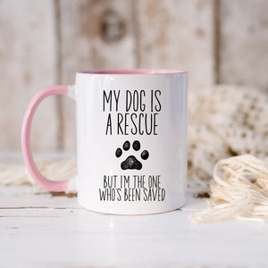 May include: White ceramic mug with a pink handle. The mug has black text that reads "My dog is a rescue but I'm the one who's been saved". A black paw print is between the two lines of text.