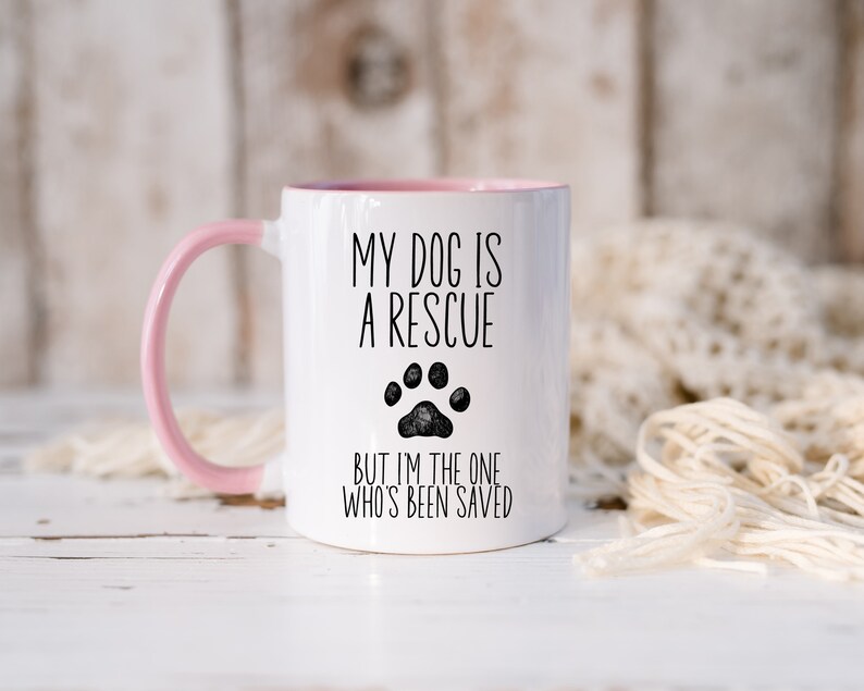 May include: White ceramic mug with a pink handle. The mug has black text that reads "My dog is a rescue but I'm the one who's been saved". A black paw print is between the two lines of text.