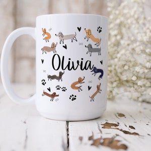 Pode incluir: Uma caneca de cer&acirc;mica branca com o nome Olivia em letra cursiva preta, rodeada por um padr&atilde;o de pequenas ilustra&ccedil;&otilde;es de c&atilde;es dachshund desenhadas &agrave; m&atilde;o em v&aacute;rios tons de castanho, cinzento e preto, com cora&ccedil;&otilde;es e pegadas pretas.