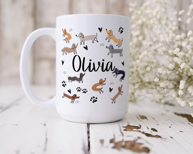 Pode incluir: Uma caneca de cer&acirc;mica branca com o nome Olivia em letra cursiva preta, rodeada por um padr&atilde;o de pequenas ilustra&ccedil;&otilde;es de c&atilde;es dachshund desenhadas &agrave; m&atilde;o em v&aacute;rios tons de castanho, cinzento e preto, com cora&ccedil;&otilde;es e pegadas pretas.