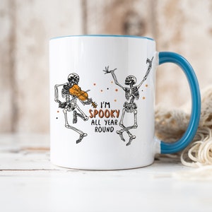 May include: White ceramic mug with a blue rim and handle. The mug features a black and white illustration of two skeletons dancing. One skeleton is playing a violin. The text on the mug reads "I'm Spooky All Year Round".