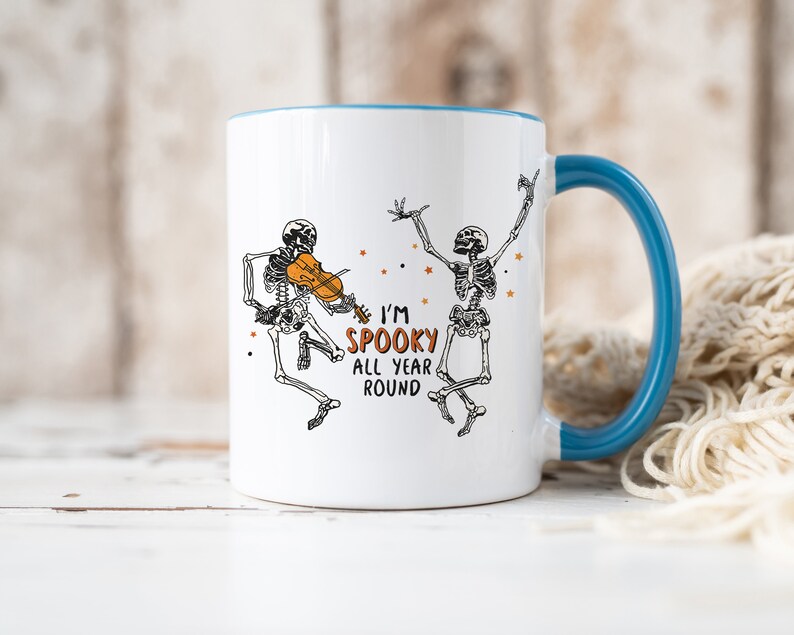 May include: White ceramic mug with a blue rim and handle. The mug features a black and white illustration of two skeletons dancing. One skeleton is playing a violin. The text on the mug reads "I'm Spooky All Year Round".
