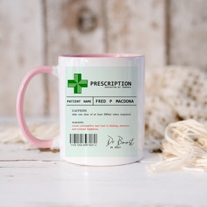 May include: A white ceramic mug with a pink handle. The mug has a green prescription label with the text "PRESCRIPTION" and "PATIENT NAME FRED P MACDONA". The label also includes the text "CAFFEINE" and "take one dose of at least 250ml when required".