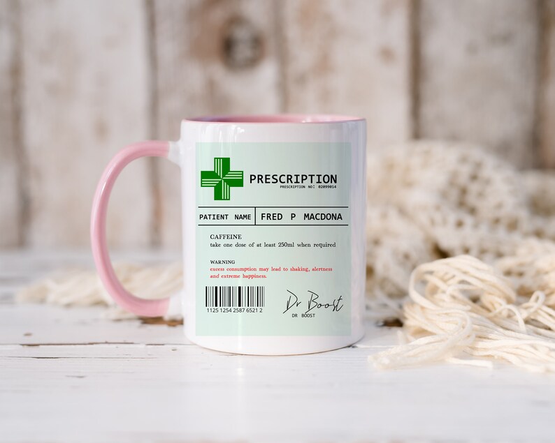 May include: A white ceramic mug with a pink handle. The mug has a green prescription label with the text "PRESCRIPTION" and "PATIENT NAME FRED P MACDONA". The label also includes the text "CAFFEINE" and "take one dose of at least 250ml when required".