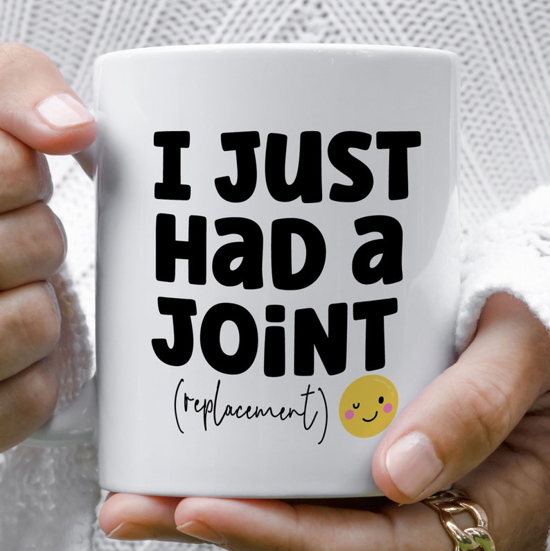 May include: A white ceramic mug featuring the black text I JUST HAD A JOINT (replacement) printed on the front. Below the text is a small yellow circular emoji with a winking face, pink cheeks, and a simple black smile.