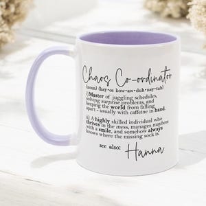 Puede incluir: Taza de cerámica blanca con borde morado. La taza presenta una definición de "Coordinador del Caos" con el nombre "Hanna" en la parte inferior.
