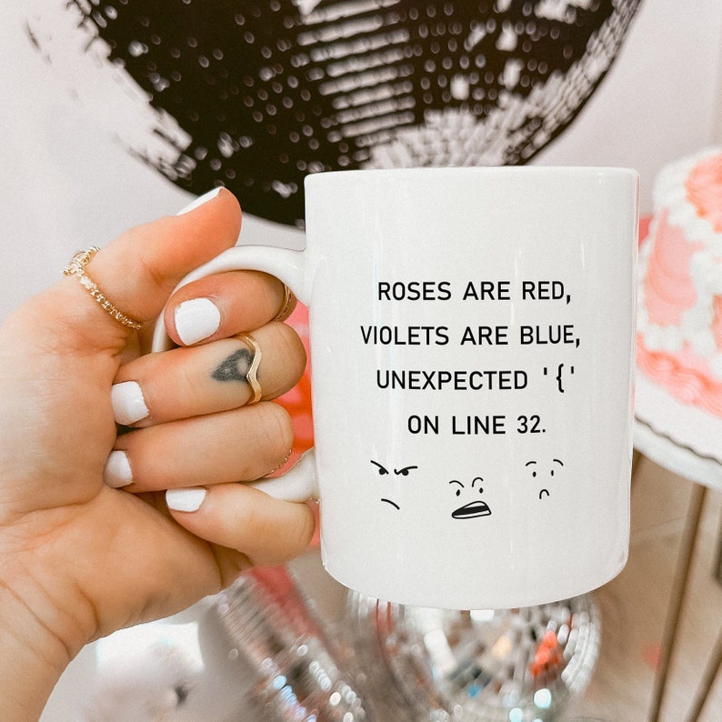 Może przedstawiać: Biały kubek ceramiczny z czarnym tekstem, kt&oacute;ry brzmi "Roses are red, violets are blue, unexpected '{' on line 32." Pod tekstem znajdują się trzy kresk&oacute;wkowe twarze, jedna zła, jedna zaskoczona i jedna smutna.