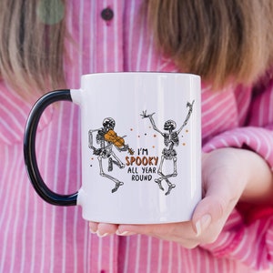 May include: White ceramic mug with a black handle. The mug features a black and white illustration of two skeletons, one playing a violin and the other dancing. The text "I'm Spooky All Year Round" is printed in black and orange on the mug.