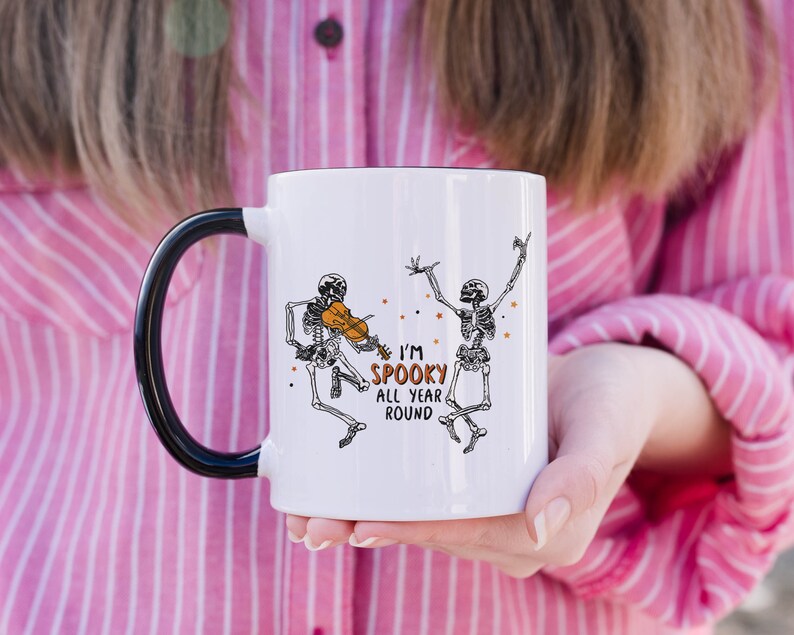 May include: White ceramic mug with a black handle. The mug features a black and white illustration of two skeletons, one playing a violin and the other dancing. The text "I'm Spooky All Year Round" is printed in black and orange on the mug.