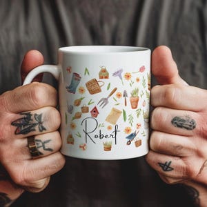 May include: White ceramic mug with a gardening theme. The mug is decorated with a variety of gardening tools, plants, and flowers. The name "Robert" is written on the mug in black cursive.