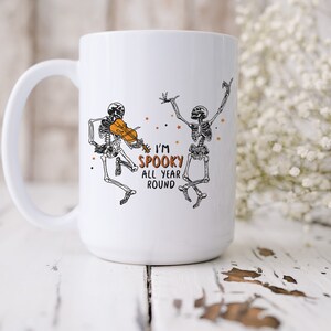 May include: White ceramic coffee mug with a black and white illustration of two skeletons. One skeleton is playing a violin and the other is dancing. The text on the mug reads "I'm Spooky All Year Round".