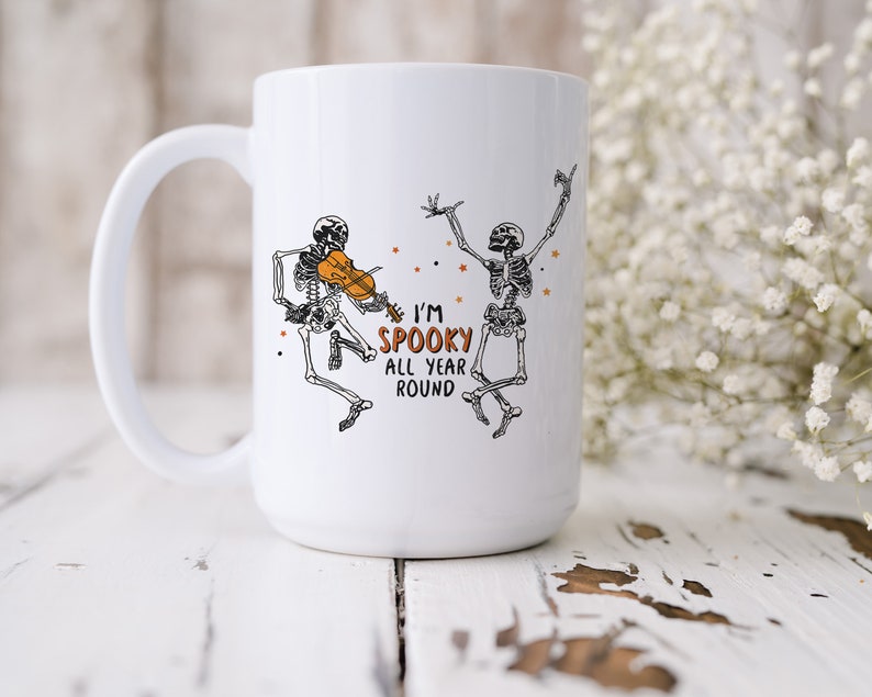 May include: White ceramic coffee mug with a black and white illustration of two skeletons. One skeleton is playing a violin and the other is dancing. The text on the mug reads "I'm Spooky All Year Round".