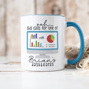 May include: A white ceramic mug with a blue handle and interior rim. The front features a graphic of a computer monitor displaying colorful bar and pie charts. The text reads: ooh... this calls for one of Brian's dashboards in black font.