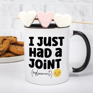 May include: A white ceramic mug with a black handle and interior rim. The front features bold black text that reads I JUST HAD A JOINT (replacement) with a small yellow smiling face icon. Three heart-shaped marshmallows are on a wooden skewer.