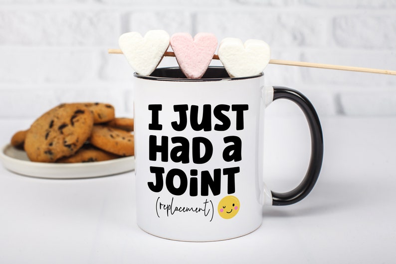 May include: A white ceramic mug with a black handle and interior rim. The front features bold black text that reads I JUST HAD A JOINT (replacement) with a small yellow smiling face icon. Three heart-shaped marshmallows are on a wooden skewer.