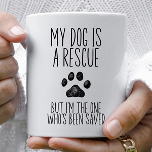 May include: White ceramic coffee mug with black text that reads "My dog is a rescue but I'm the one who's been saved". A black paw print is centered above the text.