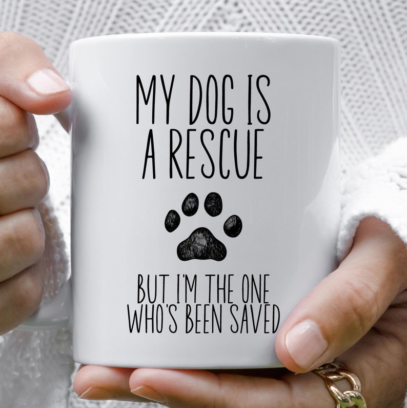 May include: White ceramic coffee mug with black text that reads "My dog is a rescue but I'm the one who's been saved". A black paw print is centered above the text.