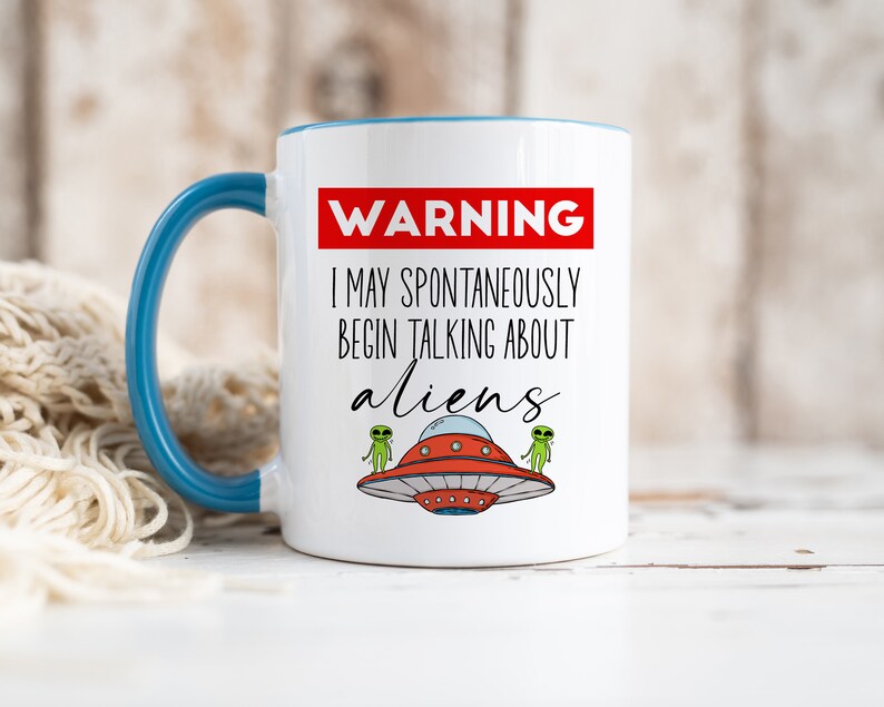 Op de afbeelding: Witte keramische mok met een blauwe rand en handvat. De mok heeft een rood waarschuwingsbord met de tekst "WAARSCHUWING IK KAN SPONTAN BEGINNEN OVER BUITENAARDSE WEZENS TE PRATEN". Er zijn twee groene buitenaardse wezens in vliegende schotels te zien.