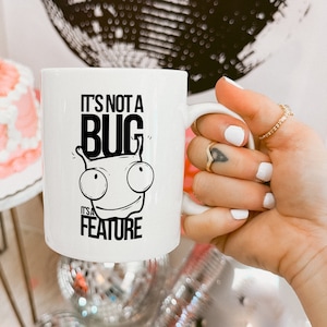 May include: White ceramic mug with a black and white illustration of a bug with the text "It's not a bug it's a feature".