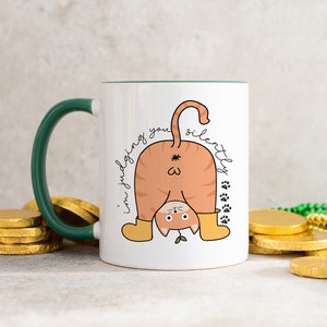 May include: White ceramic mug with a green handle. The mug features a cartoon illustration of a cat's rear end with the text "I'm judging you silently" written around it.