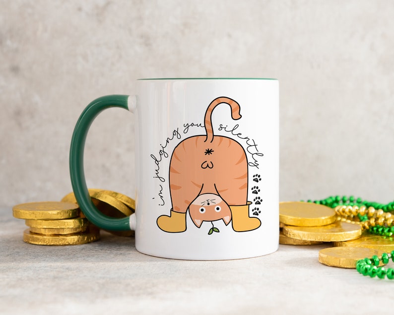 May include: White ceramic mug with a green handle. The mug features a cartoon illustration of a cat's rear end with the text "I'm judging you silently" written around it.