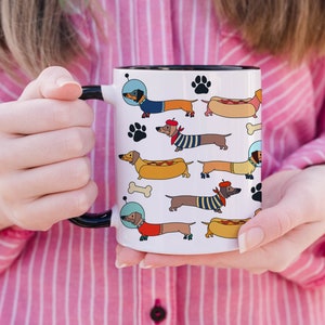 May include: White ceramic mug with a black rim and handle. The mug features a repeating pattern of cartoon dachshunds wearing various outfits, including a space suit, a beret, and a hot dog costume.
