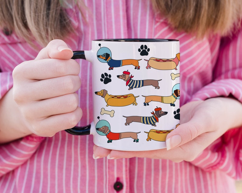 May include: White ceramic mug with a black rim and handle. The mug features a repeating pattern of cartoon dachshunds wearing various outfits, including a space suit, a beret, and a hot dog costume.