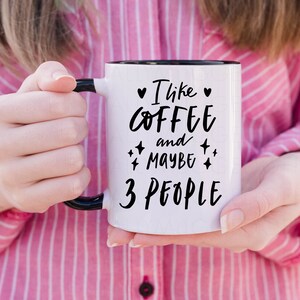 May include: White coffee mug with a black rim and handle. The mug has black text that reads "I like coffee and maybe 3 people" with hearts and stars around the text.