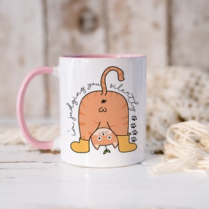 May include: White ceramic mug with a pink handle. The mug features a cartoon illustration of an orange cat with its back to the viewer, wearing yellow boots and a green plant growing from its tail. The text "I'm judging you silently" is written on the mug.