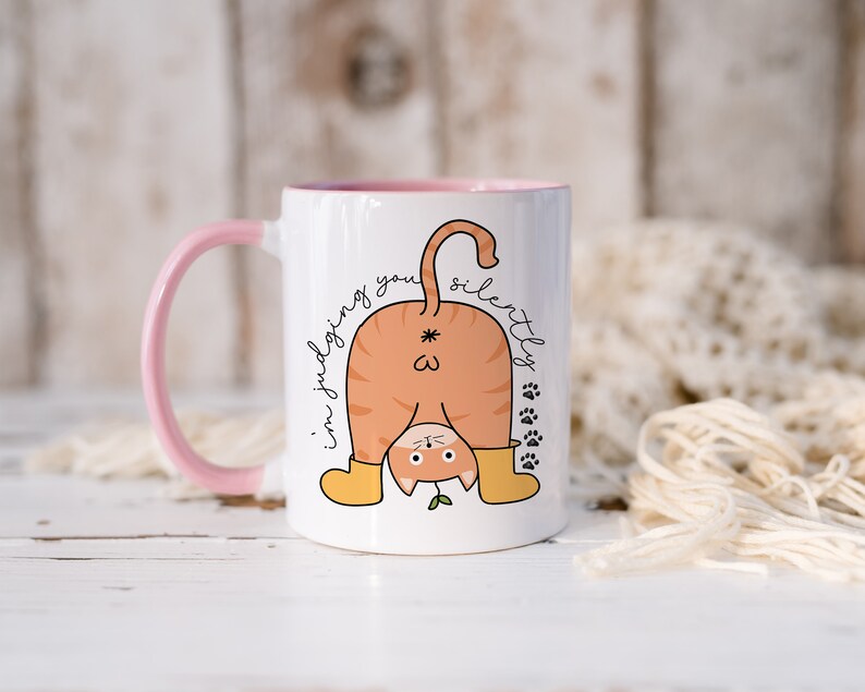 May include: White ceramic mug with a pink handle. The mug features a cartoon illustration of an orange cat with its back to the viewer, wearing yellow boots and a green plant growing from its tail. The text "I'm judging you silently" is written on the mug.