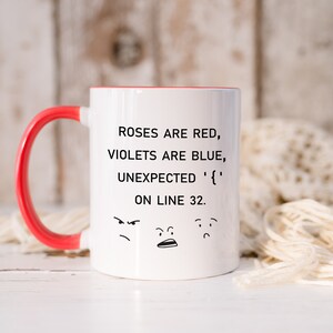 Może przedstawiać: Biały ceramiczny kubek do kawy z czerwonym uchwytem. Kubek ma czarny wz&oacute;r z tekstem "ROSES ARE RED, VIOLETS ARE BLUE, UNEXPECTED '{' ON LINE 32." i trzy kresk&oacute;wkowe twarze.