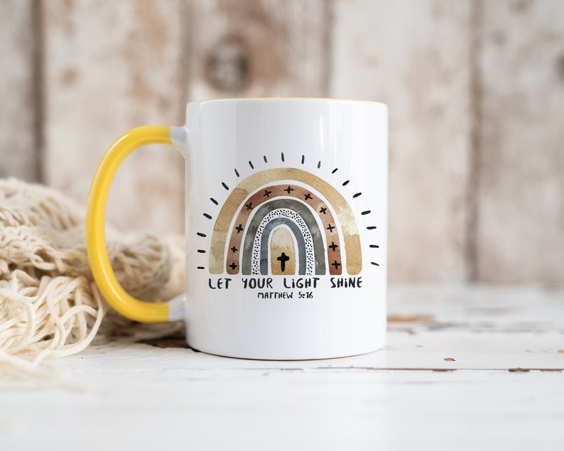 May include: White ceramic mug with a yellow handle. The mug features a watercolour rainbow with a cross in the centre and the text "Let your light shine Matthew 5:16".