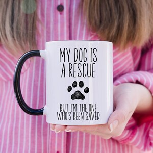 May include: White ceramic mug with a black handle. The mug has a black paw print and the text "My dog is a rescue but I'm the one who's been saved."