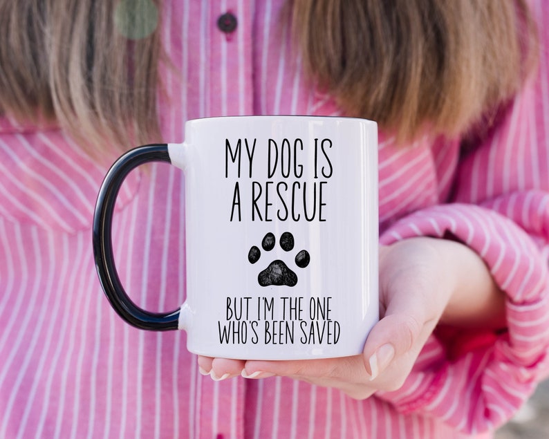 May include: White ceramic mug with a black handle. The mug has a black paw print and the text "My dog is a rescue but I'm the one who's been saved."