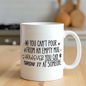 May include: White ceramic mug with black text that reads "YOU CAN'T POUR FROM AN EMPTY MUG HOWEVER YOU CAN THROW IT AT SOMEONE." The mug has a handle and is on a wooden surface.