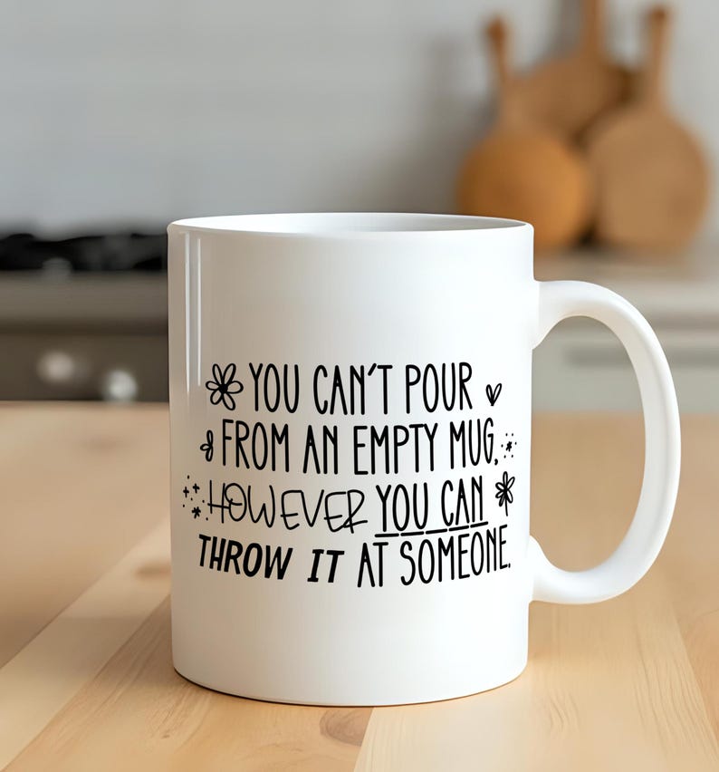 May include: White ceramic mug with black text that reads "YOU CAN'T POUR FROM AN EMPTY MUG HOWEVER YOU CAN THROW IT AT SOMEONE." The mug has a handle and is on a wooden surface.