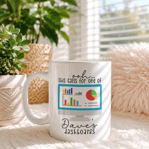 May include: A white ceramic mug featuring a graphic of a computer monitor displaying colorful bar and pie charts. The text above the monitor reads, ooh... this calls for one of, and the text below the monitor reads, Dave's dashboards.