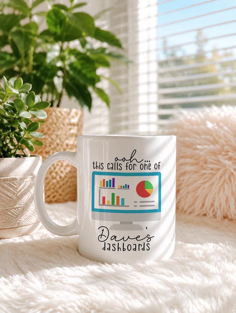 May include: A white ceramic mug featuring a graphic of a computer monitor displaying colorful bar and pie charts. The text above the monitor reads, ooh... this calls for one of, and the text below the monitor reads, Dave's dashboards.