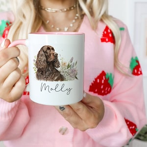 May include: White ceramic mug with a pink rim, featuring a brown and black dog with floral accents. The mug has the name "Molly" written in black cursive.
