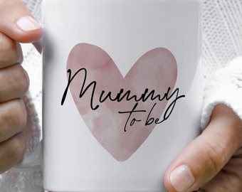 May include: A white ceramic mug featuring a large, dusty pink watercolor heart graphic centered on the front. Overlaid on the heart is the black cursive text "Mummy to be". The mug has a smooth, glossy finish and a standard cylindrical shape.