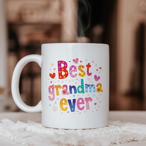 Op de afbeelding: Een witte keramische mok met de kleurrijke tekst "Best grandma ever" in een speels, meerkleurig lettertype. Het ontwerp is versierd met kleine, levendige illustraties van hartjes, sterretjes en bloemen die rond de tekst zijn verspreid.