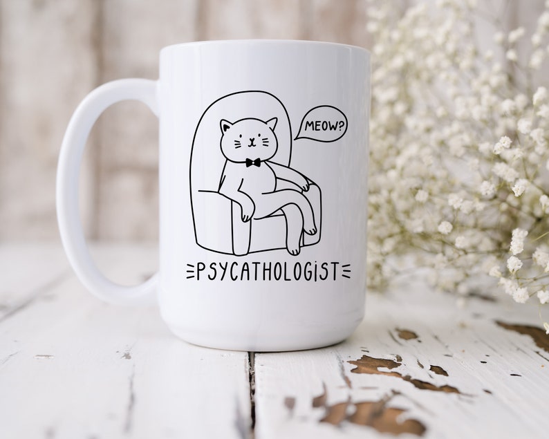 a tall white ceramic mug with an illustration of a cat, wearing a bowtie, sitting on an armchair. Beside the cat is a speech bubble with the text 'Meow?' - with text below the graphic stating 'Psycathologist'.