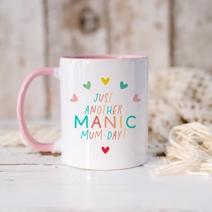 May include: A white ceramic mug with a light pink handle and interior rim. The front features the colorful text "JUST ANOTHER MANIC MUM-DAY!" surrounded by small, multi-colored hearts in shades of yellow, teal, green, and pink.
